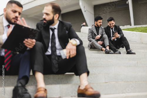 Business people outdoor meeting. A company of male businessmen in suits are sitting on the steps of the stairs. Working break. Discussion and conversation.