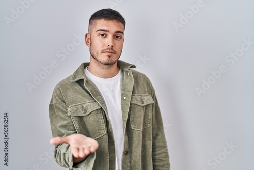 Young hispanic man standing over isolated background smiling cheerful offering palm hand giving assistance and acceptance. © Krakenimages.com