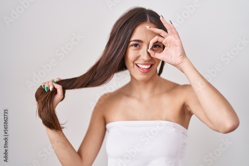 Young hispanic woman holding hair with hand smiling happy doing ok sign with hand on eye looking through fingers