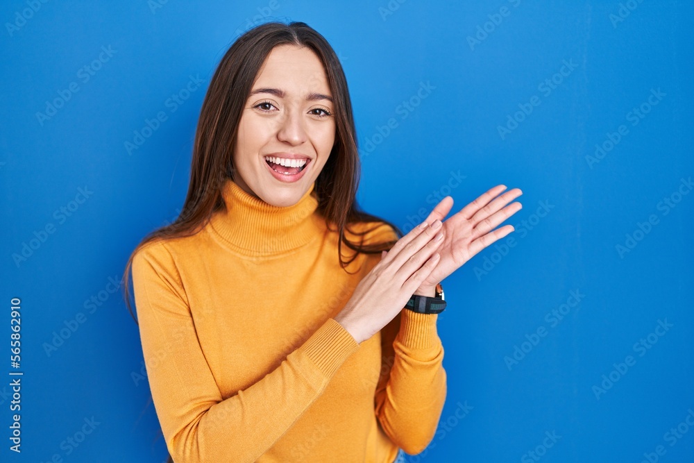 Young brunette woman standing over blue background clapping and applauding happy and joyful, smiling proud hands together