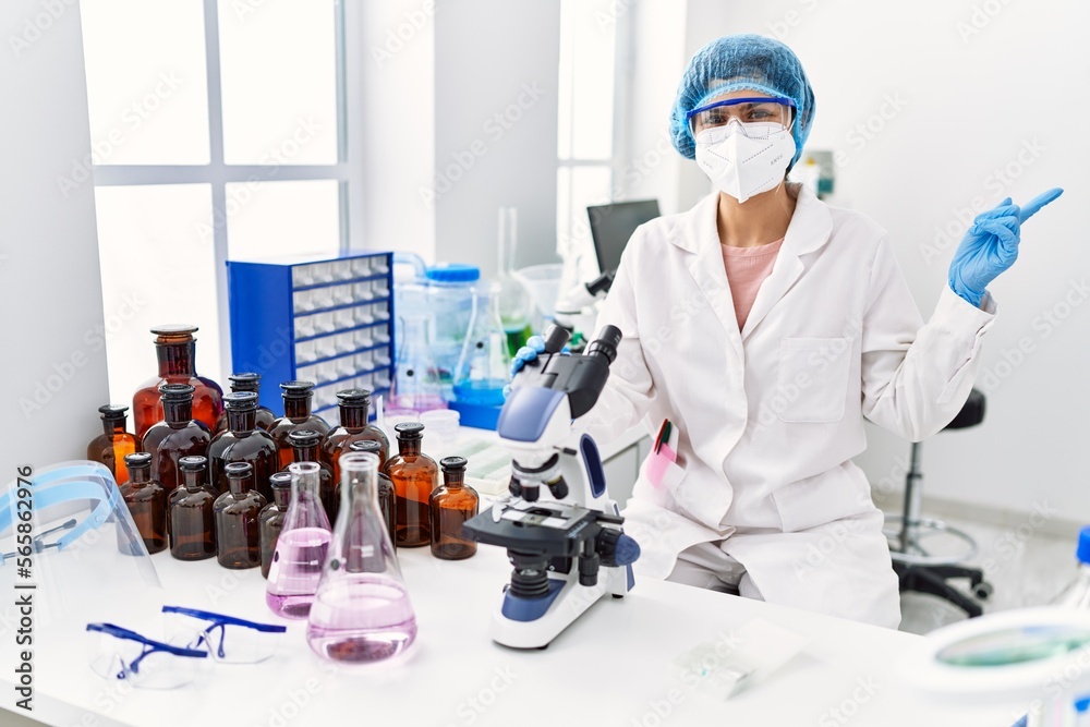 Young brunette woman working at scientist laboratory smiling happy pointing with hand and finger to the side