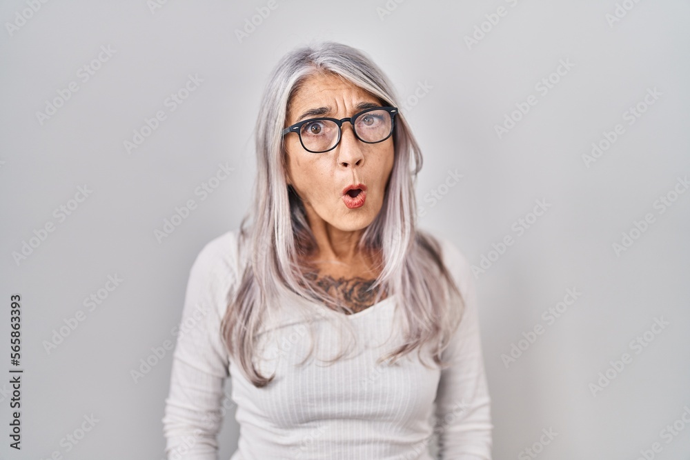 Middle age woman with grey hair standing over white background in shock face, looking skeptical and sarcastic, surprised with open mouth