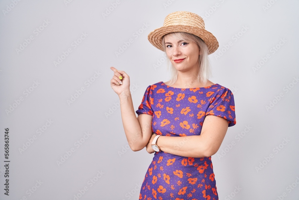 Young caucasian woman wearing flowers dress and summer hat pointing with hand finger to the side showing advertisement, serious and calm face Young caucasian woman wearing flowers dress and summer hat pointing with hand finger to the side showing advertisement, serious and calm face