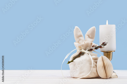 Easter bunny shaped Ivory gift bag with sweets candle and paper craft Easter egg on white wooden table, blue background banner. Willow branch