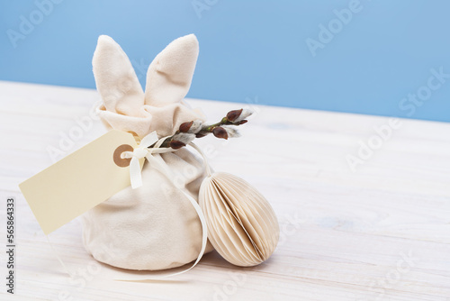 Easter bunny shaped Ivory gift bag with sweets and a mockup gift tag and paper craft Easter egg on white wooden table, blue background banner. Willow branch © taniasv