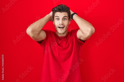 Young hispanic man standing over red background crazy and scared with hands on head, afraid and surprised of shock with open mouth