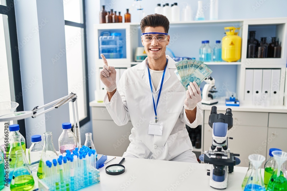 Young hispanic man working at scientist laboratory holding brazilian reals smiling happy pointing with hand and finger to the side