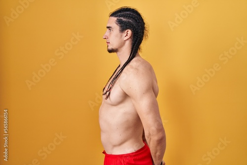 Hispanic man with long hair standing shirtless over yellow background looking to side, relax profile pose with natural face and confident smile.