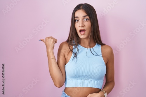 Young brunette woman standing over pink background surprised pointing with hand finger to the side  open mouth amazed expression.