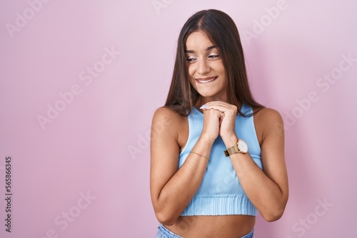 Young brunette woman standing over pink background laughing nervous and excited with hands on chin looking to the side