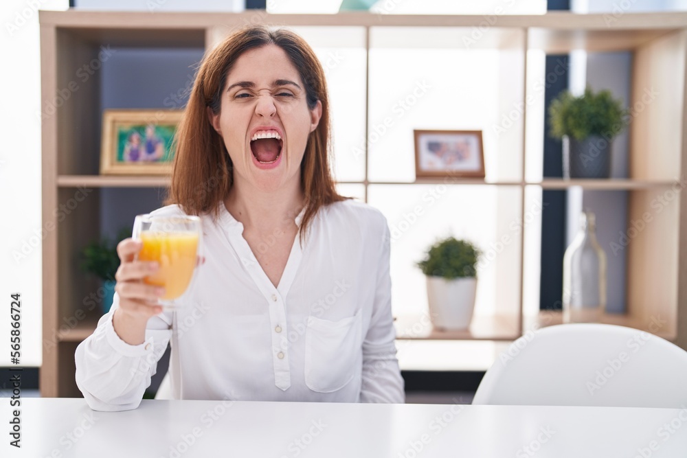 Brunette woman drinking glass of orange juice angry and mad screaming frustrated and furious, shouting with anger. rage and aggressive concept. Brunette woman drinking glass of orange juice angry and mad screaming frustrated and furious, shouting with anger. rage and aggressive concept.