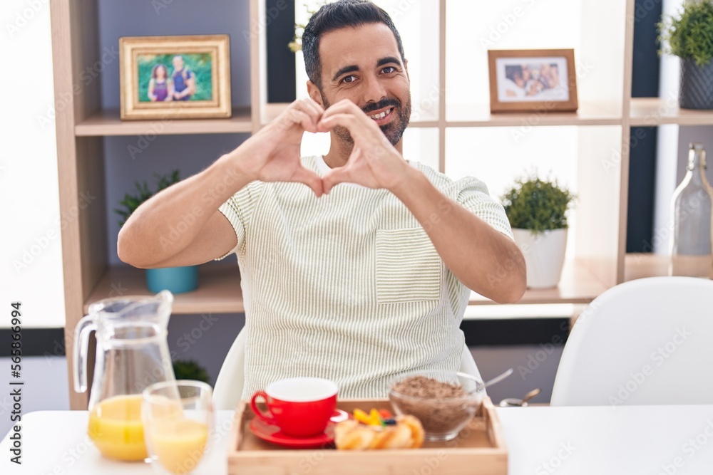 Hispanic man with beard eating breakfast smiling in love doing heart symbol shape with hands. romantic concept. Hispanic man with beard eating breakfast smiling in love doing heart symbol shape with hands. romantic concept.