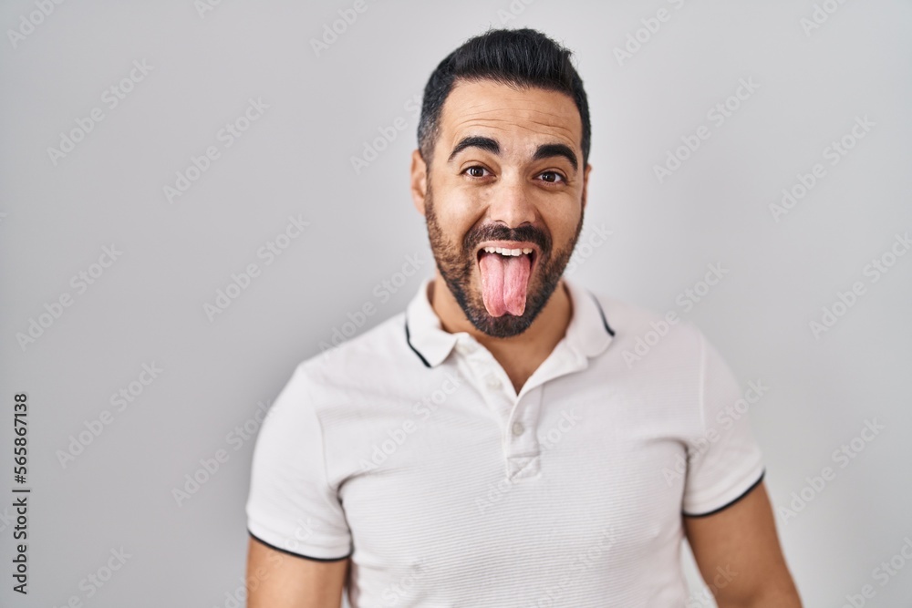 Young hispanic man with beard wearing casual clothes over white background sticking tongue out happy with funny expression. emotion concept. Young hispanic man with beard wearing casual clothes over white background sticking tongue out happy with funny expression. emotion concept.