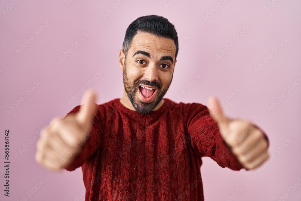 Young hispanic man with beard wearing casual sweater over pink background approving doing positive gesture with hand, thumbs up smiling and happy for success. winner gesture. Young hispanic man with beard wearing casual sweater over pink background approving doing positive gesture with hand, thumbs up smiling and happy for success. winner gesture.
