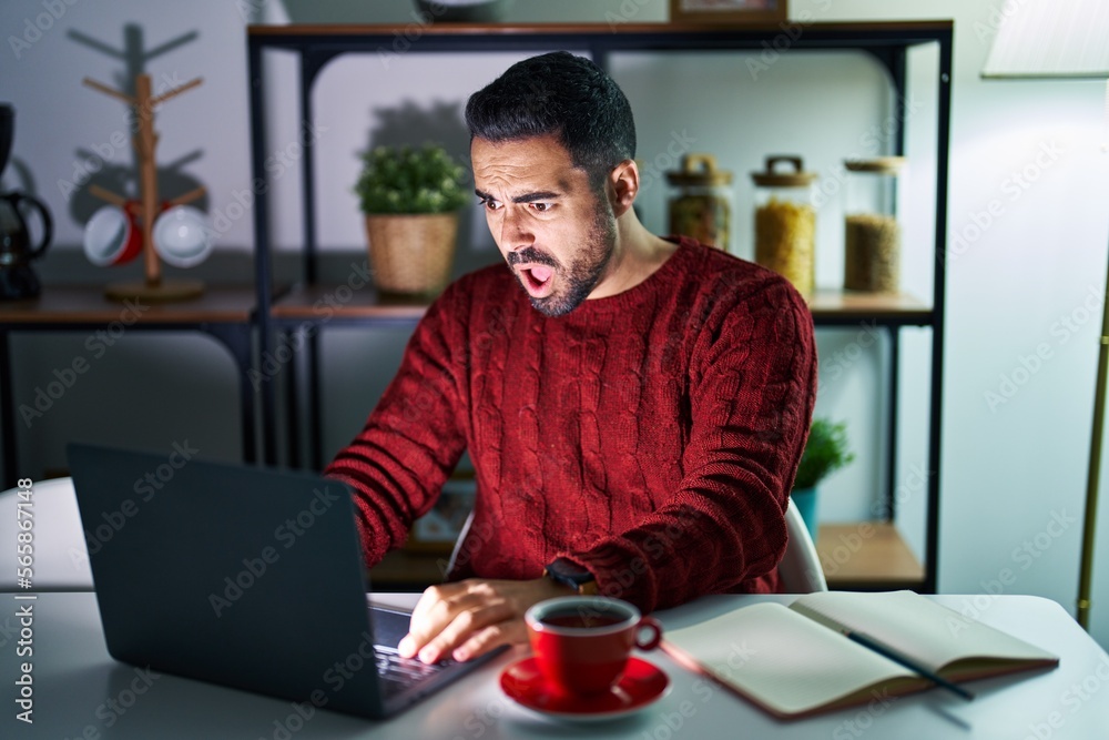 Young hispanic man with beard using computer laptop at night at home in shock face, looking skeptical and sarcastic, surprised with open mouth Young hispanic man with beard using computer laptop at night at home in shock face, looking skeptical and sarcastic, surprised with open mouth