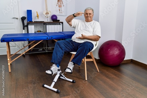 Senior caucasian man at physiotherapy clinic using pedal exerciser gesturing with hands showing big and large size sign, measure symbol. smiling looking at the camera. measuring concept.