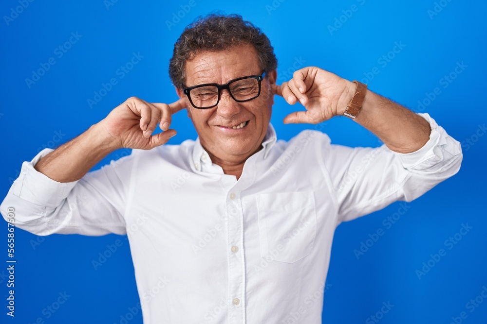 Naklejka premium Middle age hispanic man standing over blue background covering ears with fingers with annoyed expression for the noise of loud music. deaf concept.
