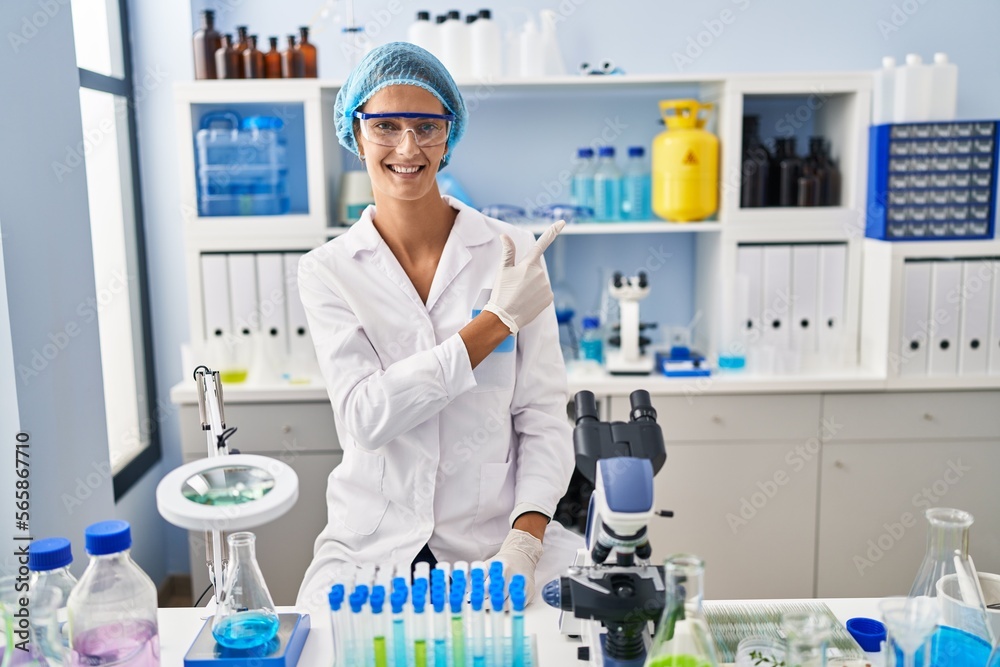 Brunette woman working at scientist laboratory cheerful with a smile on face pointing with hand and finger up to the side with happy and natural expression Brunette woman working at scientist laboratory cheerful with a smile on face pointing with hand and finger up to the side with happy and natural expression