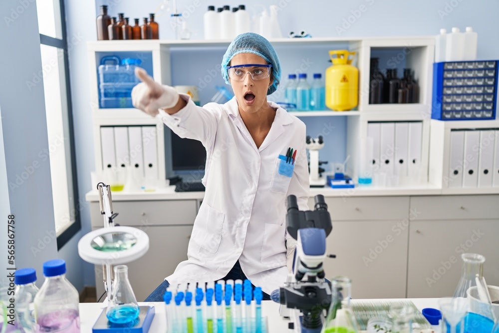 Brunette woman working at scientist laboratory pointing with finger surprised ahead, open mouth amazed expression, something on the front Brunette woman working at scientist laboratory pointing with finger surprised ahead, open mouth amazed expression, something on the front