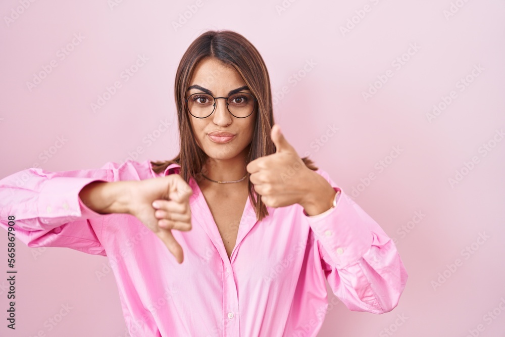 Young hispanic woman wearing glasses standing over pink background doing thumbs up and down, disagreement and agreement expression. crazy conflict Young hispanic woman wearing glasses standing over pink background doing thumbs up and down, disagreement and agreement expression. crazy conflict