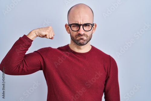 Young bald man with beard standing over white background wearing glasses strong person showing arm muscle, confident and proud of power © Krakenimages.com