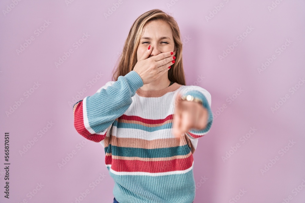 Young blonde woman standing over pink background laughing at you, pointing finger to the camera with hand over mouth, shame expression Young blonde woman standing over pink background laughing at you, pointing finger to the camera with hand over mouth, shame expression