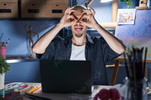 Young caucasian man using laptop at night at art studio doing ok gesture like binoculars sticking tongue out, eyes looking through fingers. crazy expression.