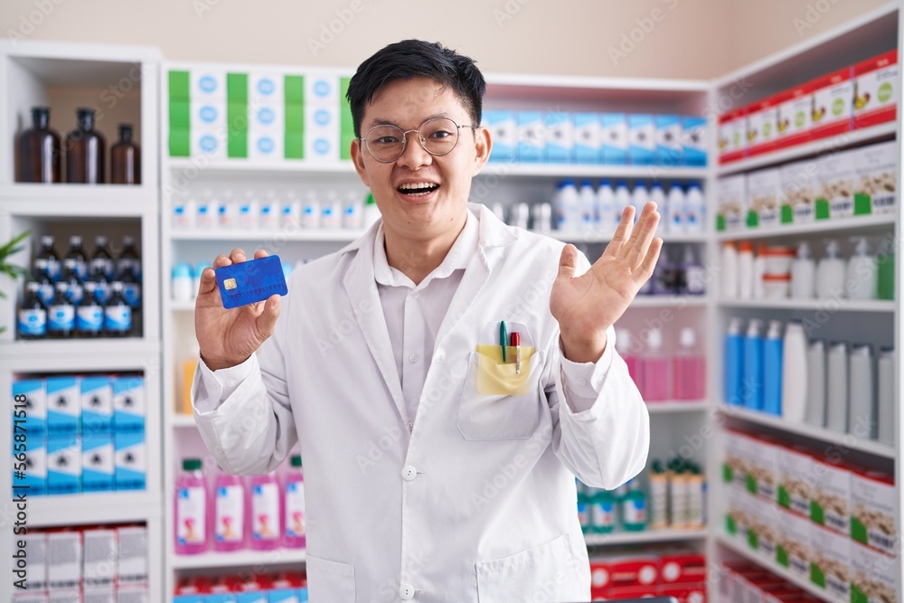 Young asian man working at pharmacy drugstore holding credit card celebrating victory with happy smile and winner expression with raised hands Young asian man working at pharmacy drugstore holding credit card celebrating victory with happy smile and winner expression with raised hands