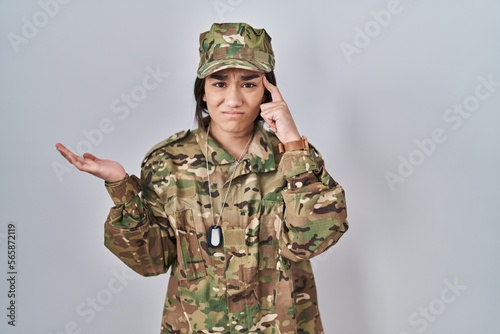 Young south asian woman wearing camouflage army uniform confused and annoyed with open palm showing copy space and pointing finger to forehead. think about it.