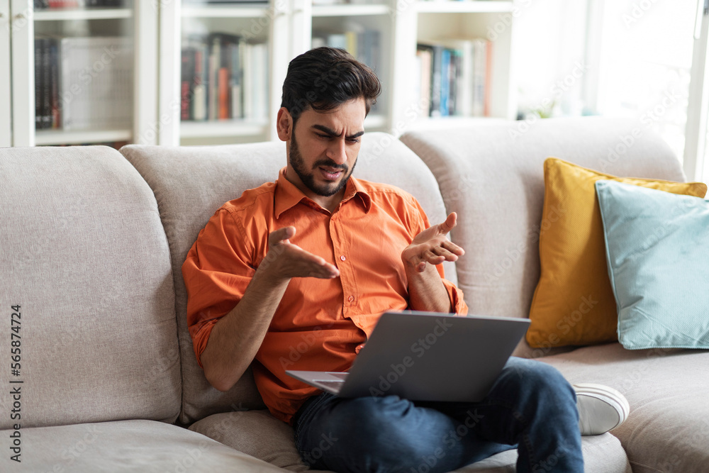 Irritated middle eastern man sitting on couch, using laptop Irritated middle eastern man sitting on couch, using laptop