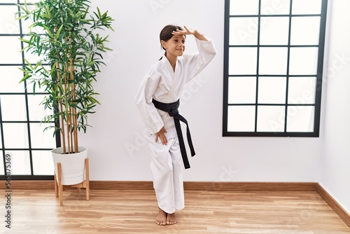 Young hispanic girl wearing karate kimono and black belt very happy and smiling looking far away with hand over head. searching concept. © Krakenimages.com