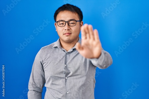 Young chinese man standing over blue background doing stop sing with palm of the hand. warning expression with negative and serious gesture on the face.
