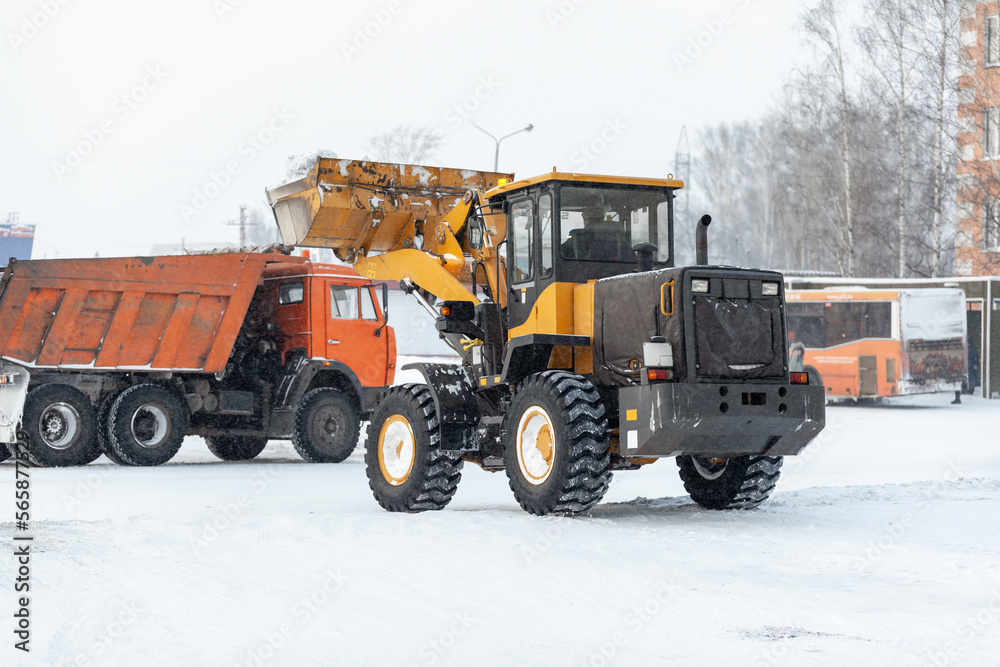 Orange tractor cleans up snow from the road and loads it into the truck. Cleaning and cleaning of roads in the city from snow in winter. Snow removal after snowfall and blizzards. Orange tractor cleans up snow from the road and loads it into the truck. Cleaning and cleaning of roads in the city from snow in winter. Snow removal after snowfall and blizzards.