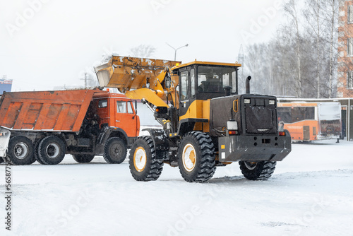 Orange tractor cleans up snow from the road and loads it into the truck. Cleaning and cleaning of roads in the city from snow in winter. Snow removal after snowfall and blizzards. 