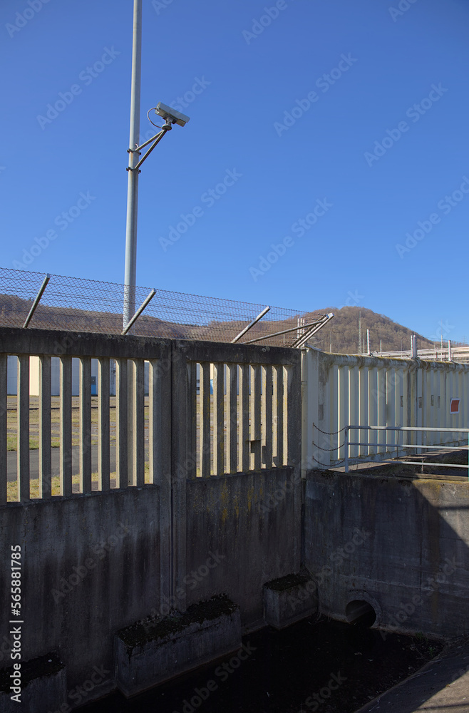 Würgassen, Germany - 03 23 2020: Security camera on a mast behind a massive concrete and steel fence Würgassen, Germany - 03 23 2020: Security camera on a mast behind a massive concrete and steel fence