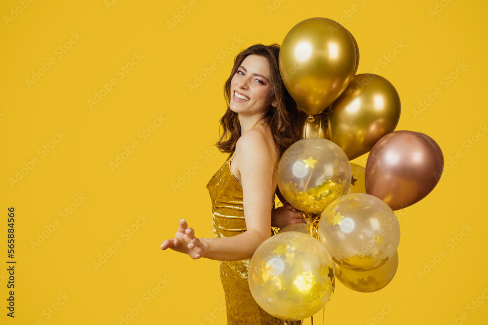 Fototapeta premium Festive concept. Charming lady in dress with bunch of air balloons having holiday party, yellow background, copy space