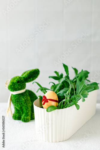 Colorful egg with a ribbon in a pot next to the green grass, next to the figure of a rabbit