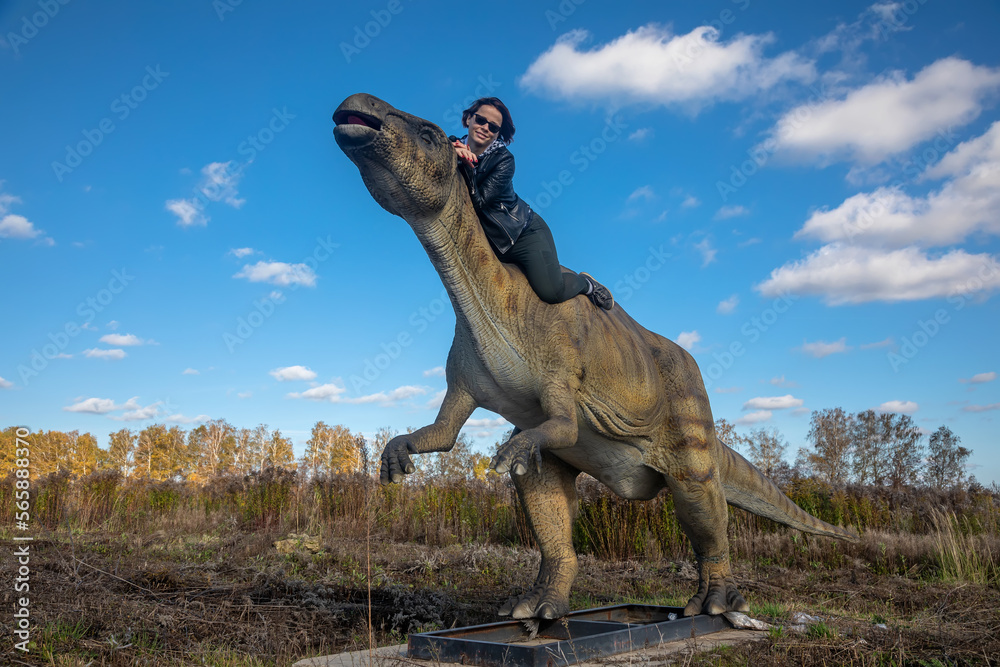Naklejka premium Beautiful girl near the dinosaur. Girl in a leather jacket. Sunny day. Dinosaur in the field.