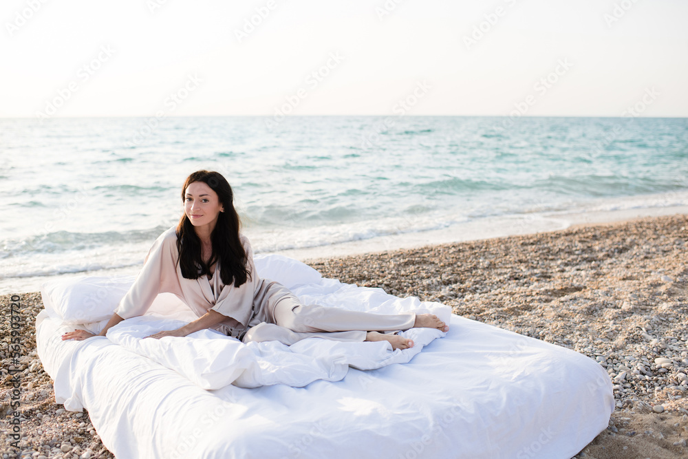Smiling young woman wear silk pajamas sitting in bed with white duvet and pillows over sea shore outdoor. Summer vacation season