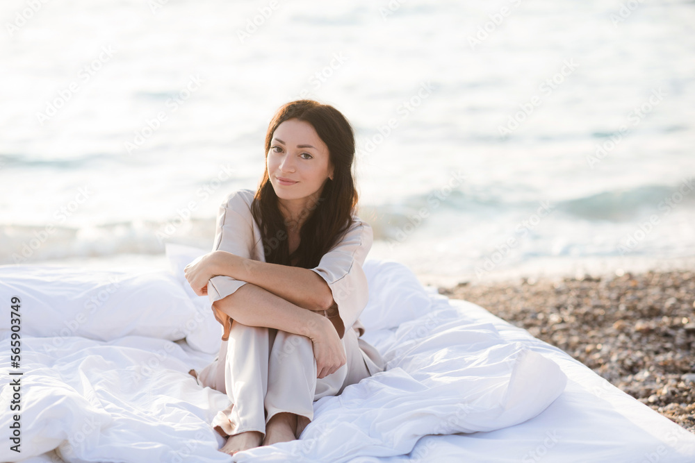 Smiling young woman wear silk pajamas sitting in bed with white duvet and pillows over sea shore outdoor. Summer vacation season