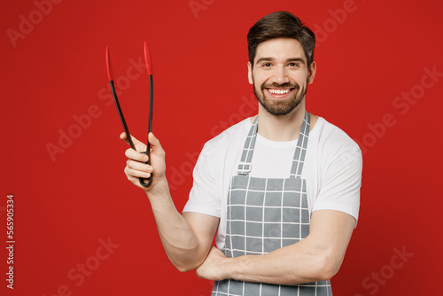 Young male housewife housekeeper chef cook baker man wear grey apron hold in hand kitchen salad serving plastic pair of tongs for grill isolated on plain red background studio. Cooking food concept. photo