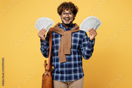 Young teen Indian boy IT student he wear casual clothes shirt glasses bag hold fan of cash money in dollar banknotes isolated on plain yellow color background. High school university college concept.