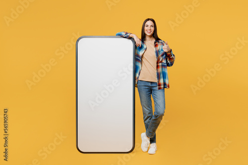Full body young woman wear blue shirt beige t-shirt big huge blank screen mobile cell phone smartphone with workspace copy space mockup area show thumb up gesture isolated on plain yellow background.