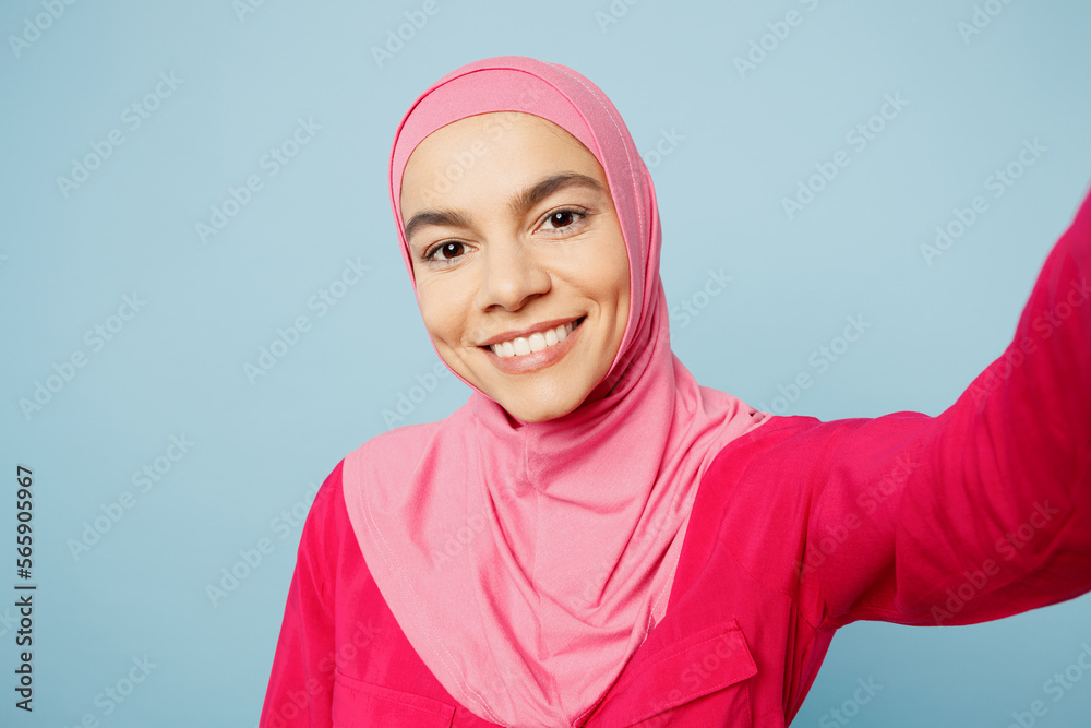 Close up fun young arabian muslim woman wear pink abaya hijab doing selfie shot pov on mobile cell phone isolated on plain pastel light blue cyan background studio. People uae islam religious concept.