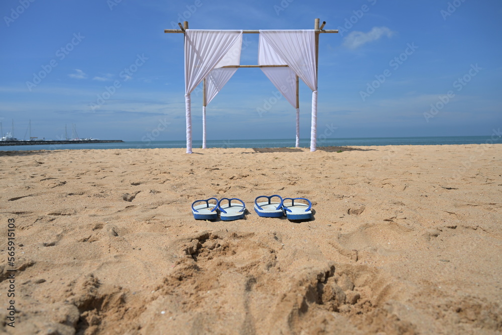 Two pairs of white flip-flops with blue ears lined up on the sandy beach fronted by bamboo arches decorated with white curtains on a summer's day.White bamboo arches installed on the beach for tourist Two pairs of white flip-flops with blue ears lined up on the sandy beach fronted by bamboo arches decorated with white curtains on a summer's day.White bamboo arches installed on the beach for tourist