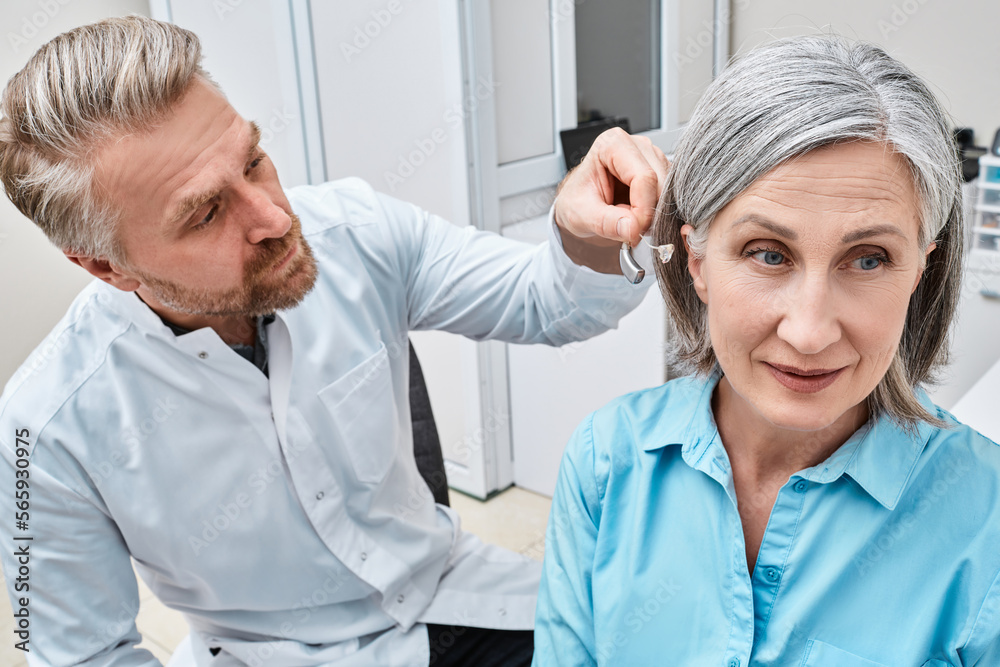 Obraz premium Audiologist fits hearing aid on deafness mature woman ear during visit to audiology center. Hearing solutions for adult