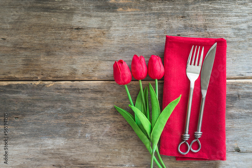 Festive place setting for romantic dinner; forged fork and knife on red napkin, bouquet of red tulips on old wooden background; space for text