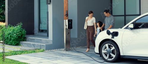 Fotografia Progressive young parents and daughter with electric vehicle and home charging station