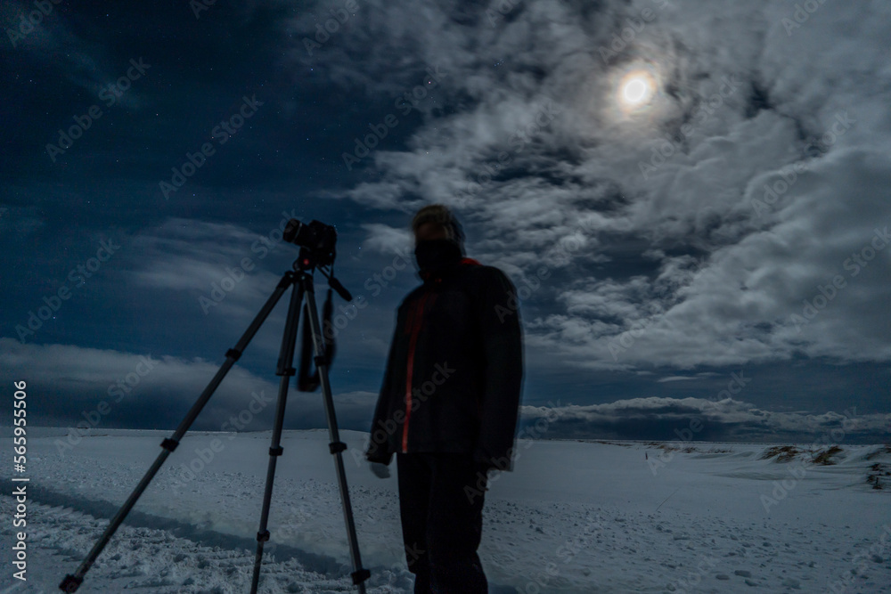 Silhouette of a photographer, camera and tripod on a totally snowy esplanade with the full moon illuminating the cloudy and star-filled sky at night in Iceland
