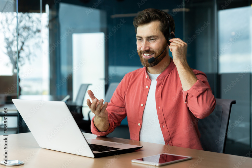 Portrait of a young man in a headset and a red shirt. He sits at the desk in the office, works on a laptop, communicates through a video call, talks, smiles.